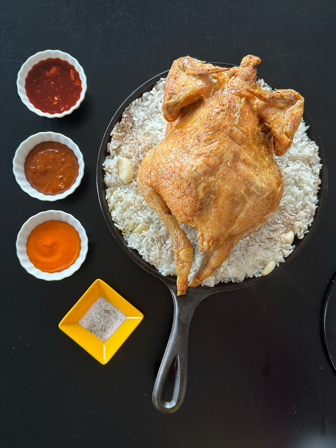 Fried chicken on rice with three dipping sauces in a cast iron skillet on a black surface.